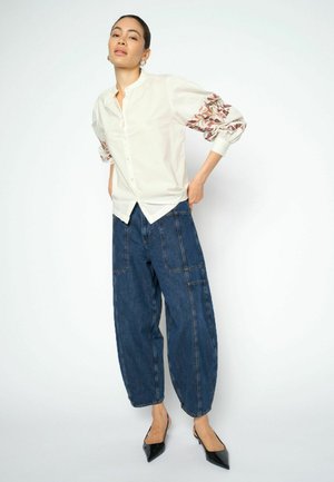 Donna che indossa una camicetta bianca con ricami floreali sulle maniche, jeans blu larghi e tacchi neri appuntiti, in piedi su sfondo bianco.