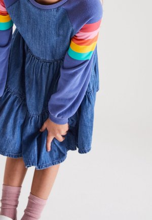 Enfant portant une robe en jean et un chemisier à manches longues rayé de couleurs vives, soulevant légèrement la robe d'une main.