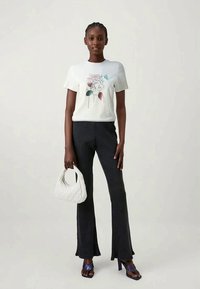 Mulher vestindo uma t-shirt gráfica branca com flores, calças pretas à boca de sino, sandálias roxas de salto alto, segurando uma mala branca texturizada contra um fundo simples.
