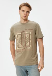 Beige bomulds t-shirt med et centralt maske grafik i brunt og creme, med dekorative mønstre og teksten "VÆR MODIG" ovenfor.