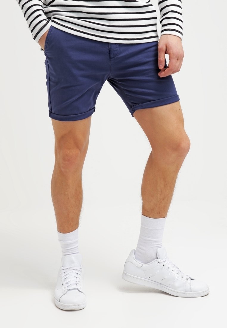 Homme portant un short bleu marine retroussé, des chaussettes blanches, des baskets blanches et un haut à manches longues rayé noir et blanc, les mains dans les poches.