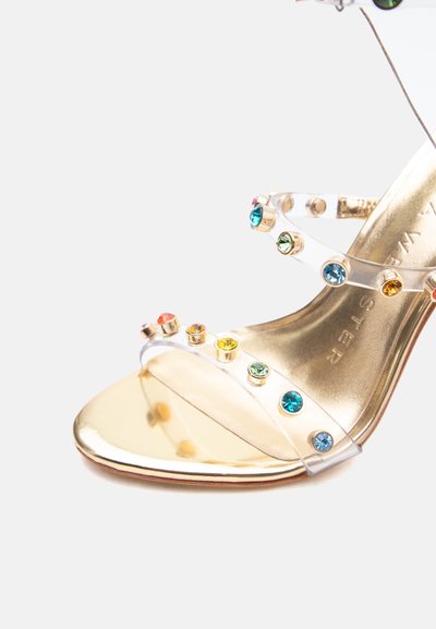Χρυσό σανδάλι με ψηλό τακούνι, διαφανείς λουρίδες διακοσμημένες με πολύχρωμους στρογγυλούς πολύτιμους λίθους και λουρί στον αστράγαλο με πόρπη.