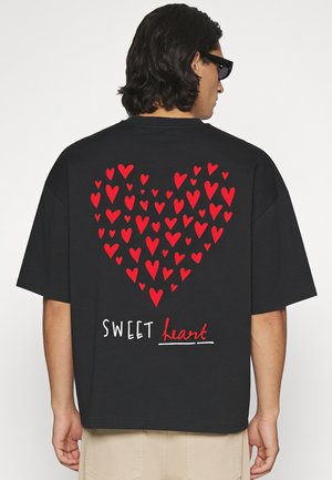 Homme portant un t-shirt noir avec un cœur rouge composé de petits cœurs et le texte "SWEET heart" dans le dos, pantalon beige et lunettes de soleil noires.