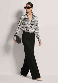 Schwarz-weiß gemustertes Button-Up-Shirt, hoch taillierte schwarze Nadelstreifen-Hosen und cremefarbene spitze High Heels. Die Accessoires umfassen eine schwarze Clutch und Sonnenbrille.