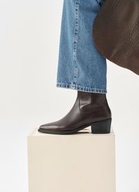 Stivale alla caviglia in pelle marrone con punta affilata, tacco basso a blocco e pannelli laterali elastici, abbinato a jeans blu a gamba corta.