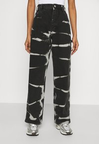 Pantalones de mezclilla anchos de color negro con un patrón de tie-dye blanco, cintura alta, cierre de un botón y bolsillos laterales. Combinados con zapatillas deportivas plateadas.