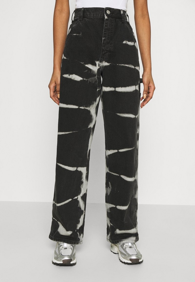 Pantalones de mezclilla anchos de color negro con un patrón de tie-dye blanco, cintura alta, cierre de un botón y bolsillos laterales. Combinados con zapatillas deportivas plateadas.