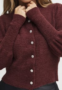 Burgunderfarbener Strickcardigan mit langen Ärmeln, ausgestattet mit großen Kristallknöpfen und gerippten Bündchen. Weiche Textur mit leicht verkürztem Design.