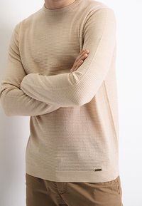 Homme portant un pull beige texturé à manches longues, bras croisés, associé à un pantalon marron sur un fond blanc uni.