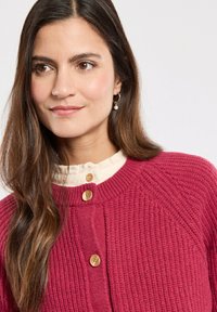 Cardigan en tricot bordeaux avec un motif texturé, présentant un col rond et des boutons en bois. Porté par-dessus un chemisier de couleur crème.