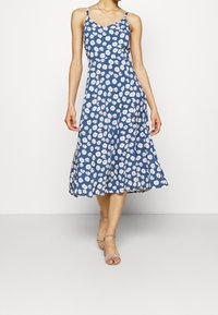 Robe fleurie bleue avec motif de marguerites blanches, sans manches, et longueur midi évasée. Tissu doux avec une texture subtile et décolleté en V.