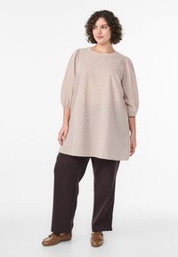 Lekka beżowa tunika w wzory z bufkami, połączona z ciemnobrązowymi, luźnymi spodniami i brązowymi butami na sznurówki.