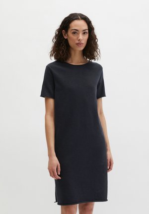 Kobieta o kręconych włosach, ubrana w czarną, jednolitą sukienkę do kolan z krótkim rękawem, stojąca na białym tle.