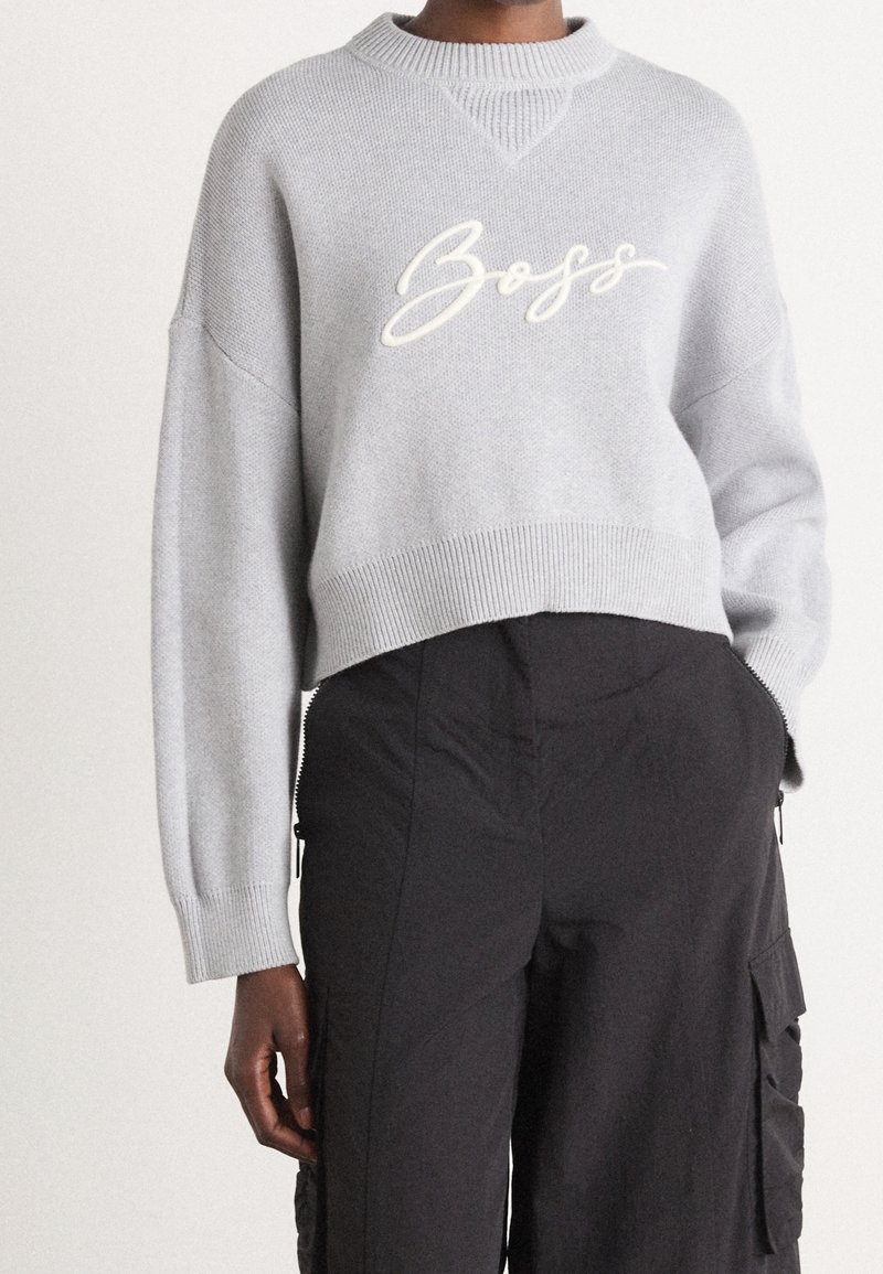 Pull court gris avec le texte « Boss » associé à un pantalon cargo noir taille haute avec poches latérales, porté par une personne debout.