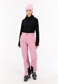 Jonge vrouw in roze skibroek, zwarte coltrui, roze muts en zwarte skischoenen staand tegen een witte achtergrond.