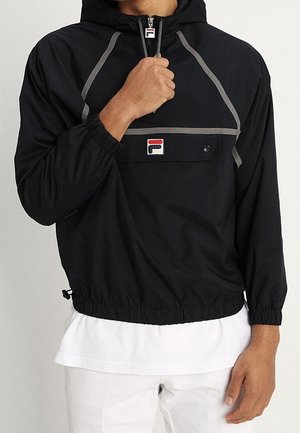 Persona che indossa una felpa nera Fila con bordo grigio e patch con logo, parzialmente aperta, sopra una camicia bianca e pantaloni bianchi.