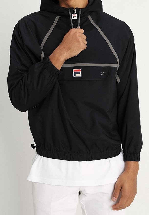 Personne portant un sweat à capuche noir Fila avec un liseré gris et un patch logo, partiellement zippé, sur une chemise blanche et un pantalon blanc.