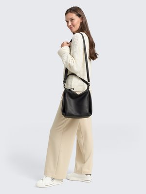 Femme en pull crème et pantalon beige portant un sac à bandoulière en cuir noir, souriant et regardant par-dessus son épaule sur un fond gris clair.