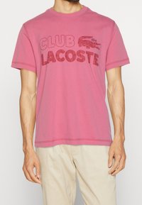 Man med en rosa kortärmad T-shirt med "CLUB LACOSTE" och en krokodil-logotyp i rött på framsidan, ihop med beige byxor.