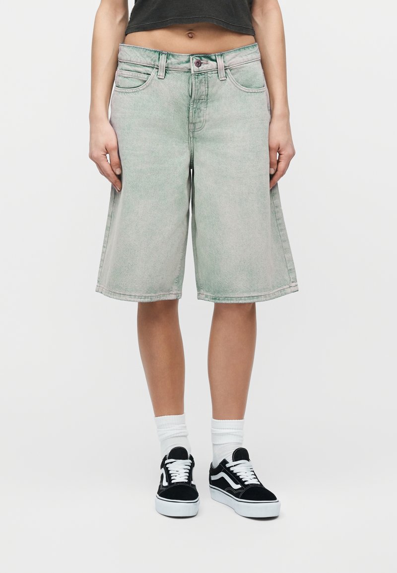 Άτομο φοράει ανοιχτό πράσινο τζιν σορτς μέχρι το γόνατο, μαύρο κοντό τοπ, λευκές κάλτσες και μαύρα-άσπρα αθλητικά παπούτσια, στέκεται μπροστά σε απλό φόντο.