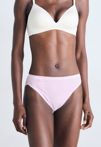 Femme à la peau foncée portant un soutien-gorge crème et des culottes en coton rose clair, se tenant debout avec les bras détendus le long du corps sur un fond clair et uni.