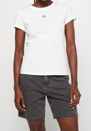 Femme portant un t-shirt blanc à manches courtes avec un petit logo et un short en jean noir taille haute avec des bords effilochés.