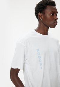 Vit bomullströja med vertikal blå text "NOBODY." Korta ärmar med klassisk rund halsringning. Slät textur.