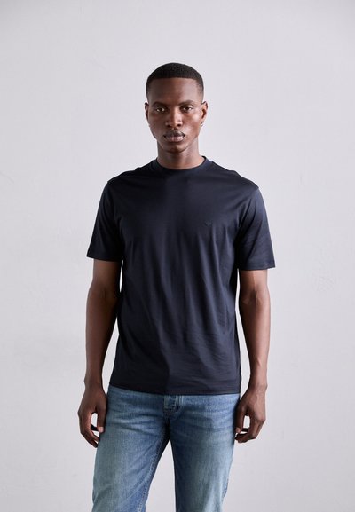 Mørkeblå kortærmet t-shirt lavet af glat bomuld, med rund hals og et diskret logo på brystet. Båret med lyseblå jeans.