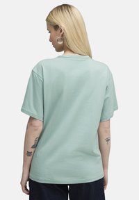 Miętowy bawełniany t-shirt z krótkim rękawem, okrągłym dekoltem i luźnym krojem. Widoczne tatuaże na ramieniu. Prosty widok z tyłu.