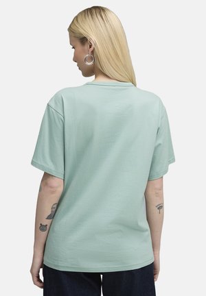 Camiseta de algodón verde menta con mangas cortas, escote redondo y un corte relajado. Tatuajes visibles en el brazo. Vista trasera simple.