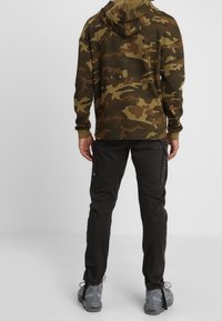Hoodie w kamuflażowym wzorze w ziemistych odcieniach, zestawiony z czarnymi spodniami cargo. Obuwie modela ma szare akcenty i sznurowadła, widoczne na dole.