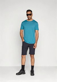 Pávatollzöld rövid ujjú ing, sötét rövidnadrág, fekete zokni és fekete cipő. A modell sima háttér előtt áll, napszemüveget visel.