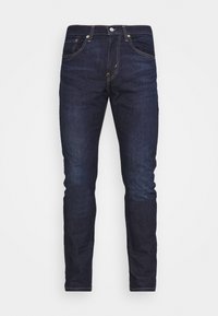 Tmavě modré slim-fit džíny s předními a zadními kapsami a viditelným oranžovým švem na bílém pozadí.