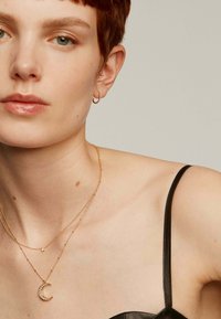 Joyería en tono dorado con collares en capas que presentan un colgante de luna creciente. La modelo lleva una blusa de cuero negra. Cabello corto y recortado.