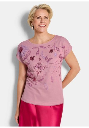 Lächelnde blonde Frau, die ein rosa floral besticktes Oberteil und einen leuchtend pinken Satinhrock trägt, vor einem weißen Hintergrund stehend.