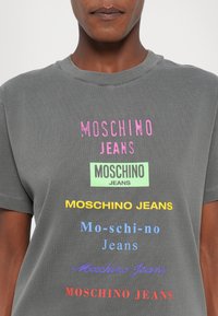 Grå bomullstrøye med flere fargerike trykk av "Moschino Jeans" i forskjellige fonter og farger, inkludert rosa, grønn, gul og blå.
