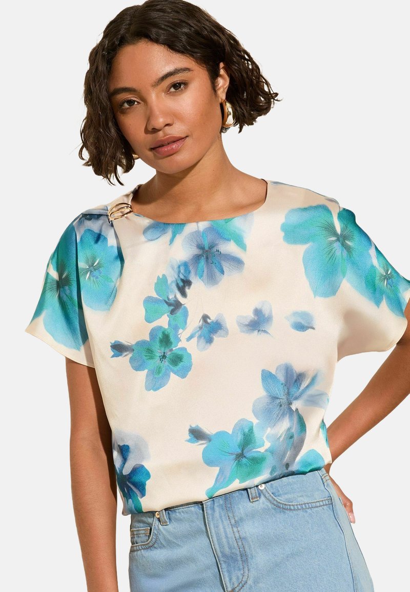 Blommig blus i lätt tyg med blå blommor på en krämvit bakgrund, båtutskuret halslinningar, korta ärmar och en distinkt detalj i guld.