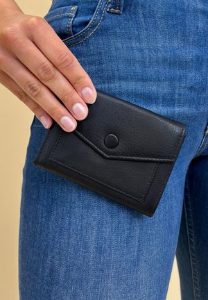 Black leather wallet with a textured finish, featuring a triangular flap closure and a snap button, held against blue denim jeans.