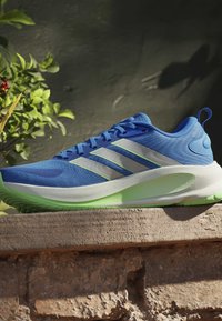 Chaussure de course bleue avec semelle blanche et vert citron reposant sur un rebord en pierre avec un feuillage vert en arrière-plan.