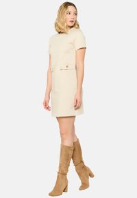 Beige Kurzarmkleid mit tailliertem Oberteil, eingeschnürtem Bund und einer Taschenverzierung auf der Vorderseite. Kombiniert mit beigen knielangen Wildlederstiefeln.