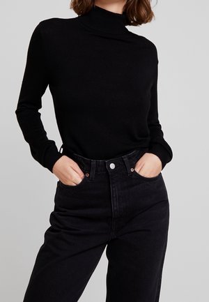 Femme portant un pull à col roulé noir à manches longues rentré dans un jean noir taille haute, mains dans les poches, sur un fond gris clair.
