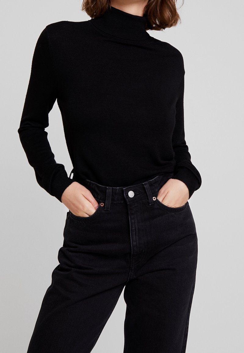Femme portant un pull à col roulé noir à manches longues rentré dans un jean noir taille haute, mains dans les poches, sur un fond gris clair.