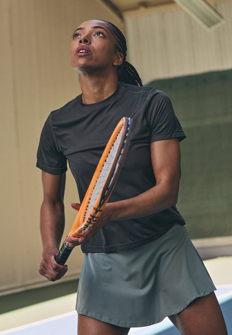 Camisa de deporte negra de manga corta y falda verde clara llevadas por una persona que sostiene una raqueta de tenis naranja, mirando hacia arriba contra un fondo neutro.