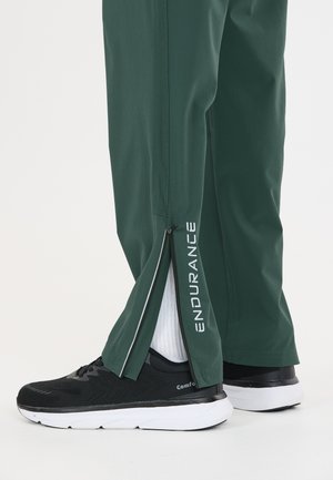 Pantalon vert avec fermeture éclair dézippée à la cheville révélant des chaussettes blanches et des chaussures de sport noires avec des semelles blanches, texte "ENDURANCE" sur la jambe du pantalon.