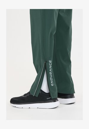 Pantalon vert avec fermeture éclair dézippée à la cheville révélant des chaussettes blanches et des chaussures de sport noires avec des semelles blanches, texte "ENDURANCE" sur la jambe du pantalon.