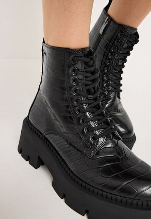 Bottes noires à cheville avec une texture crocodile, dotées d'un design à lacets, d'une semelle en caoutchouc épaisse et de coutures angulaires.