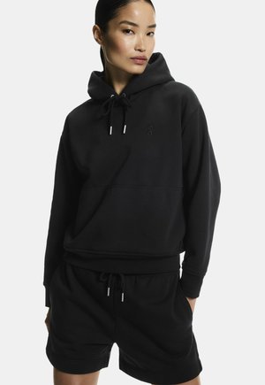 Femme portant un sweat à capuche noir avec cordons et un short noir assorti, debout les mains dans les poches devant un fond clair uni.
