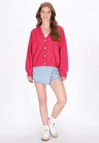 Rosa gestrickte Strickjacke mit V-Ausschnitt, Knopfverschluss und metallischen Fäden, kombiniert mit einem Jeans-Minirock und weißen Sneakern.