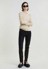 Γυναίκα φορώντας μαύρα γυαλιά ηλίου, μπεζ ριγέ πουλόβερ, μαύρα εφαρμοστά τζιν και μαύρες πλατφόρμες, στέκεται με τα χέρια σταυρωμένα.