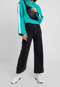 Haut à capuche court turquoise avec des rayures blanches sur les côtés, associé à un jean large noir et des baskets blanches. Sac en bandoulière noir porté en travers du corps.